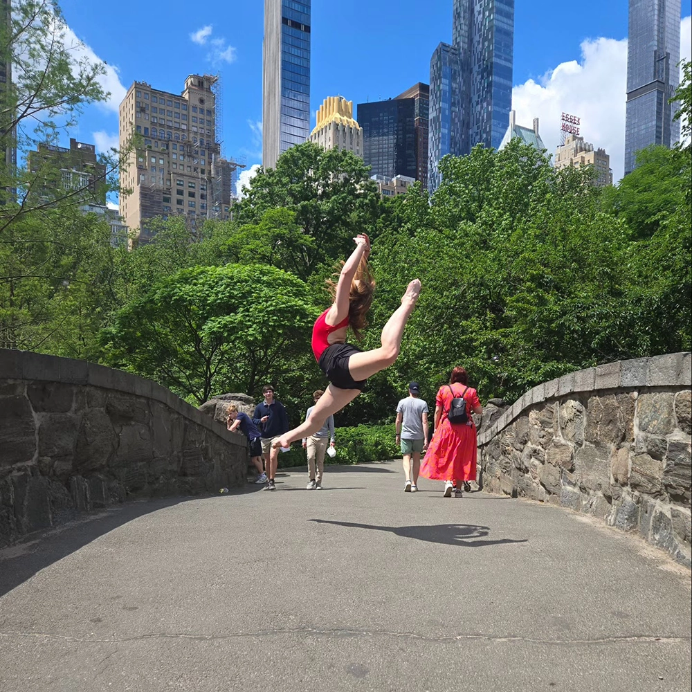 A young woman wearing a short, black ballet skirt and a red leotard leaps into the air in an arabesque, hands raised, on a bridge in Central Park. Big green trees and the skyscrapers of NYC are in the background. A few other pedestrians are also on the bridge. 