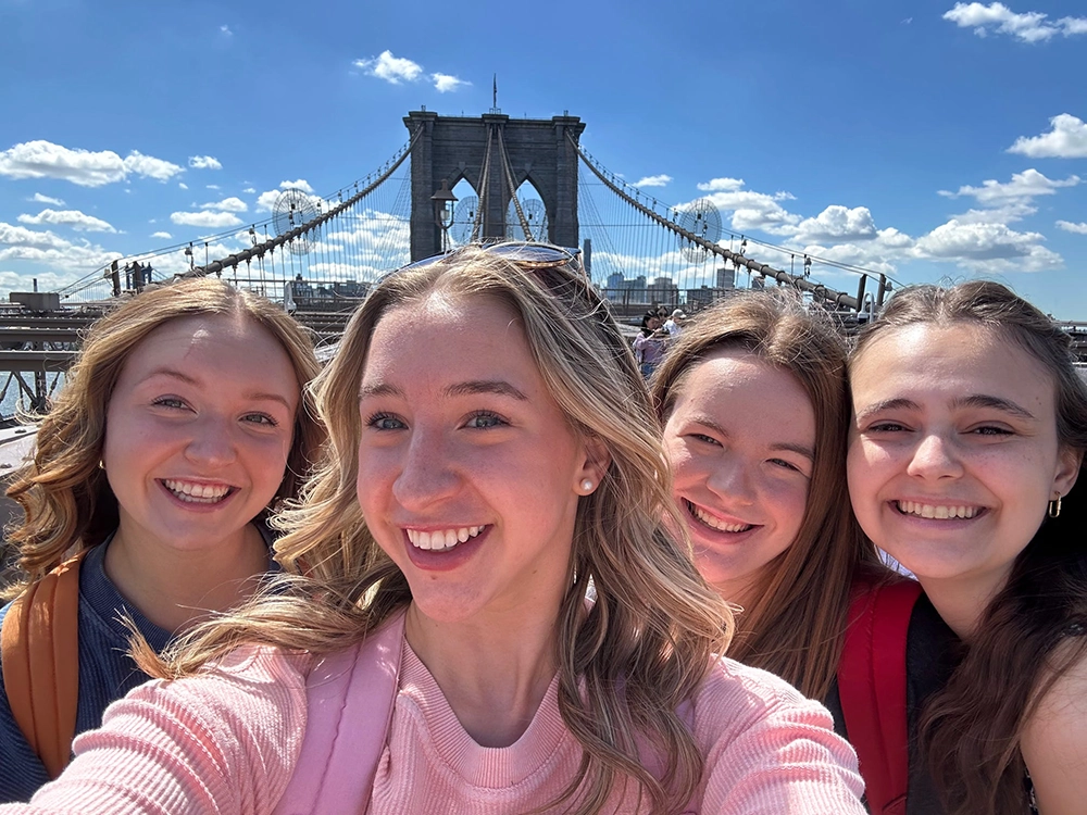 Four young women, all with long hair and big smiles, pose together in a selfie on the Brooklyn Bridge. 