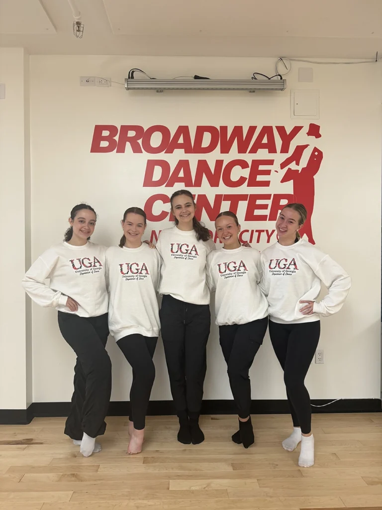 Five young women, all wearing black pants and white sweatshirts that read UGA Department of Dance, post in front of a white wall with "Broadway Dance Center New York City" written in red.