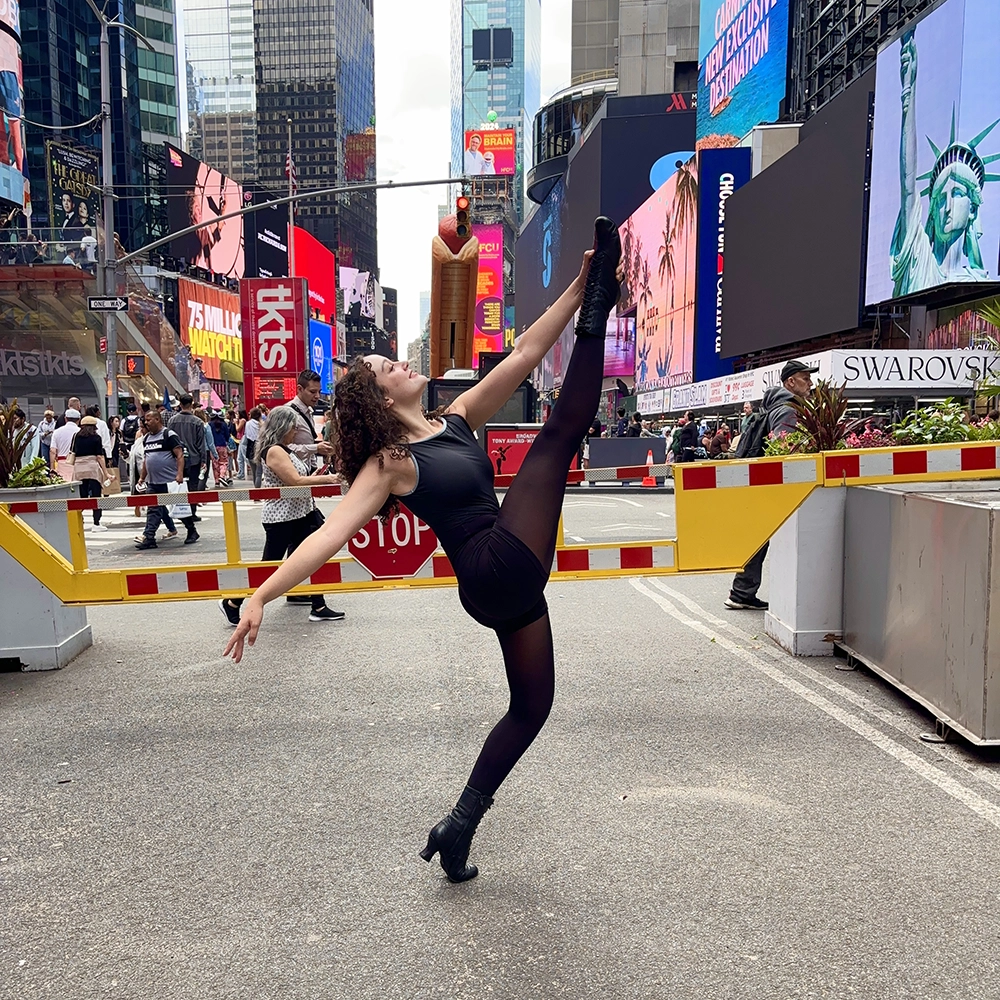 A young woman dressed all in black holds one leg straight up in the air on a city street. One hand holds the instep of the lifted foot, and the other is stretched out behind her. The skyscrapers and video screens of Times Square in NYC are in the background. 