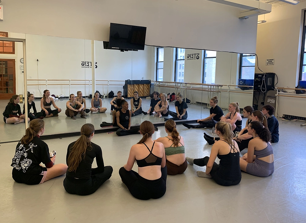 In a dance studio with a mirrored wall and barres pushed against the sides of the room, a dance instructor who is sitting cross legged on the floor, talks to a group of dance students who are also on the floor. The students are gathered in a half circle and listening intently. 