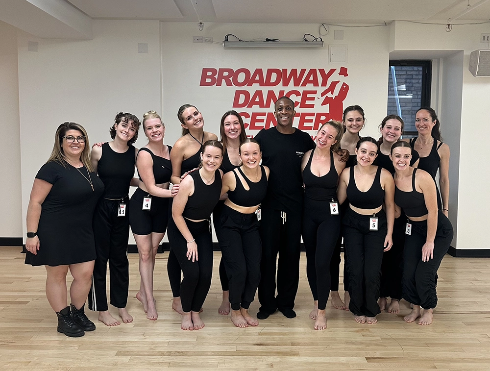 A group of dance students all dressed in black leotards and tights stand in a group for a photo with two dance instructors at Broadway Dance Center in NYC.