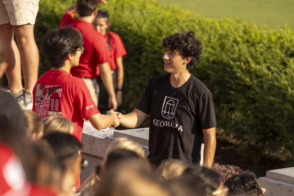 College student JJ Lazo shakes hands with another college student. He is wearing a black university shirt while the other student is wearing a red shirt.