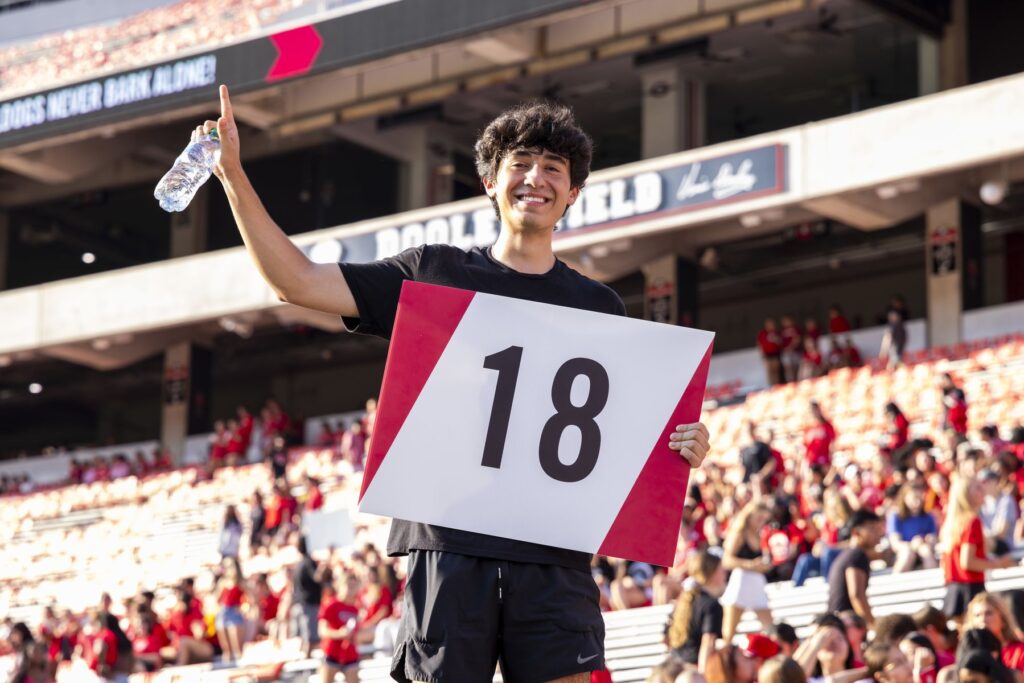 College student JJ Lazo stands up in the bleachers at a UGA football game. He is wearing black clothing and holding a sign with the number 18 on it.