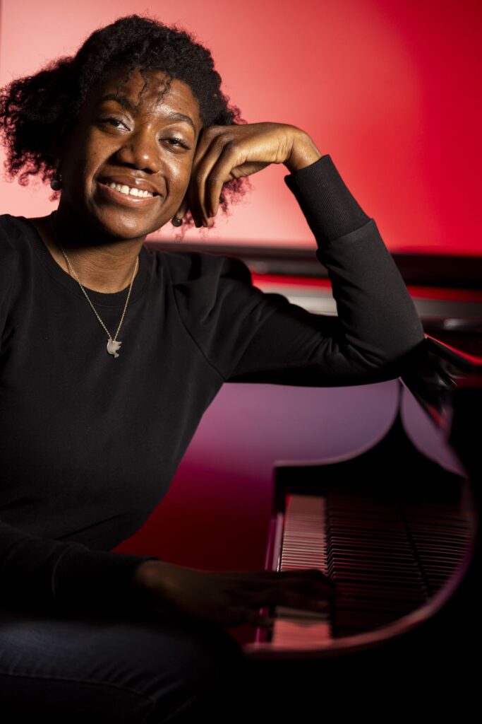 A photo of Asia Simone Passmore sitting at a piano. She is backlit by a gentle red light and smiling as she rests an elbow on the piano.