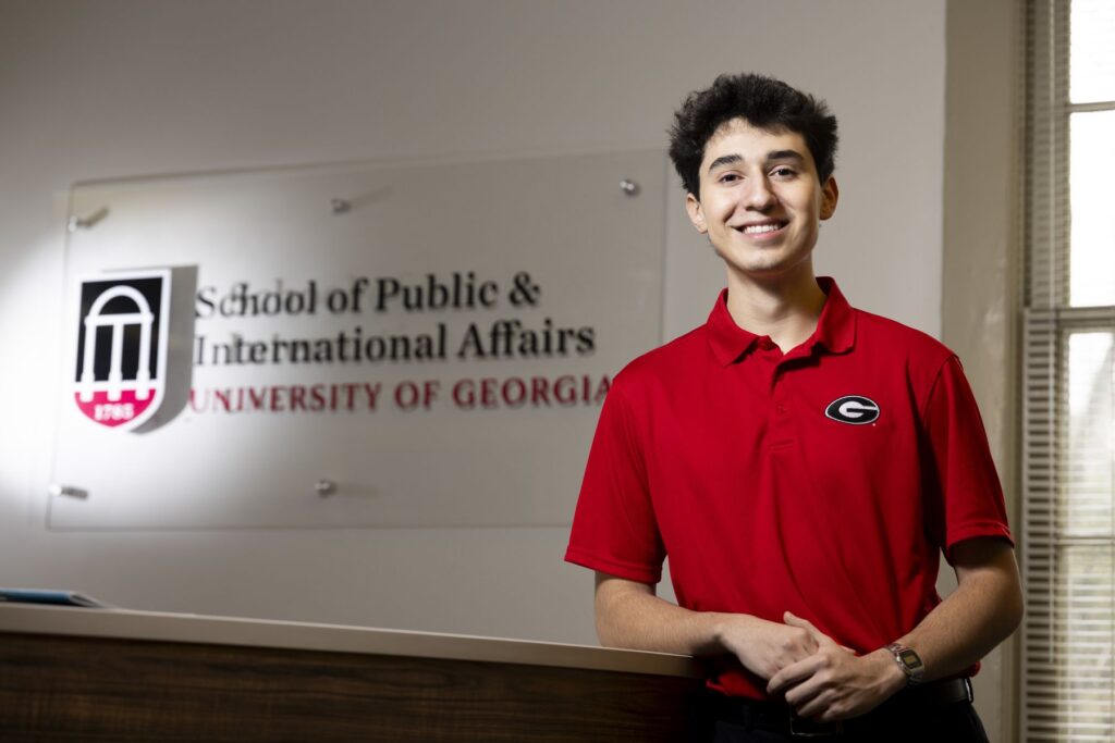 College student JJ Lazo poses in front of a University logo. He is wearing a red UGA shirt and smiling at the camera.