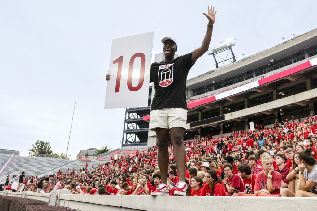 UGA student Stephen Amolegbe stands on the barrier of a stadium with the stadium crowd seated behind him. Most of the crowd is dressed in red. Stephen holds a white sign with the number 10 in red on it. He is wearing a black UGA-branded shirt with white shorts.