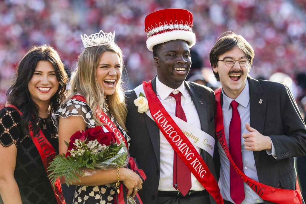Four members of UGA's homecoming court pose as a blurred stadium crowd cheers behind them. The homecoming court members are wearing formal wear and red sashes.