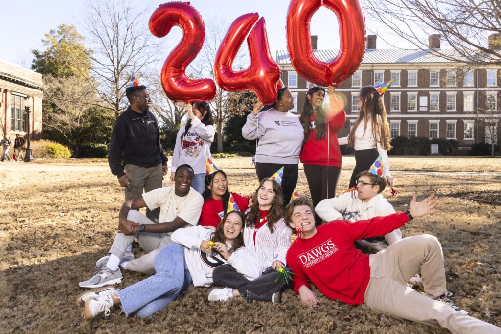 Several UGA students pose on the grass outside of a university building. They are holding three large red balloons shaped like a