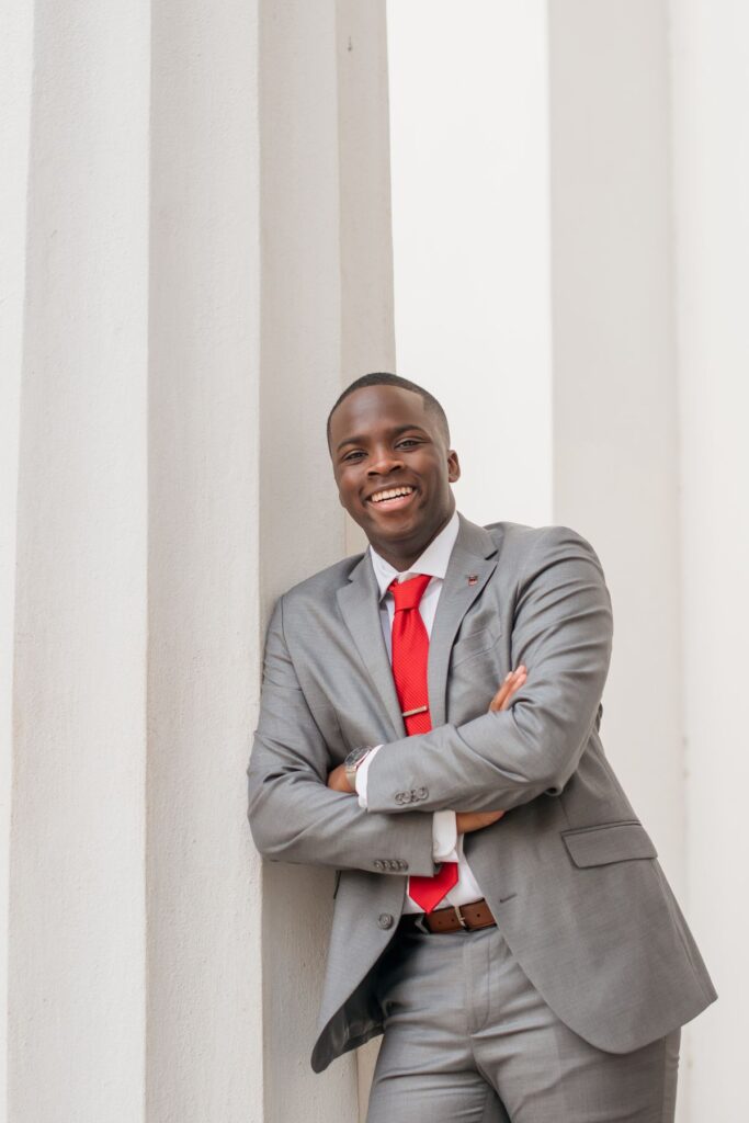 Stephen Amolegbe, a UGA student, poses against tall, white columns. He is wearing a grey suit with a red tie. His arms are crossed.