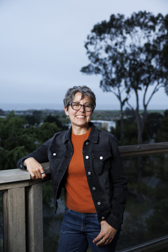 A women wearing a jean jacket, jeans, and an orange t-shirt stands outside on a wooden deck. There are blurred green trees behind her.
