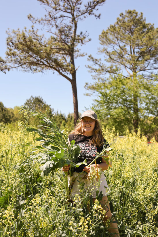 A young woman stands in a field holding a large bunch of greenery.
