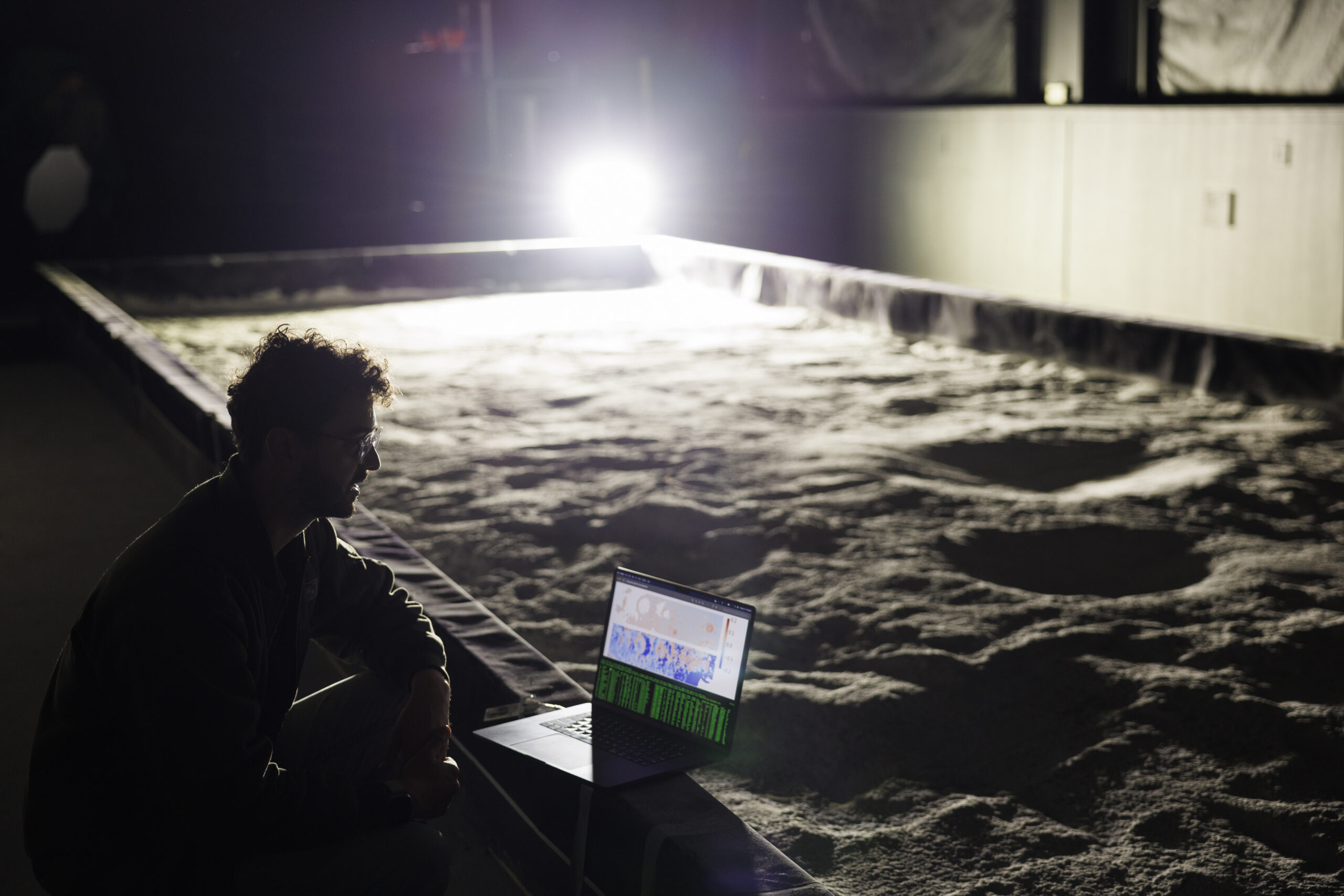 A darkened image of a man with a computer observing a plot of sand indoors.