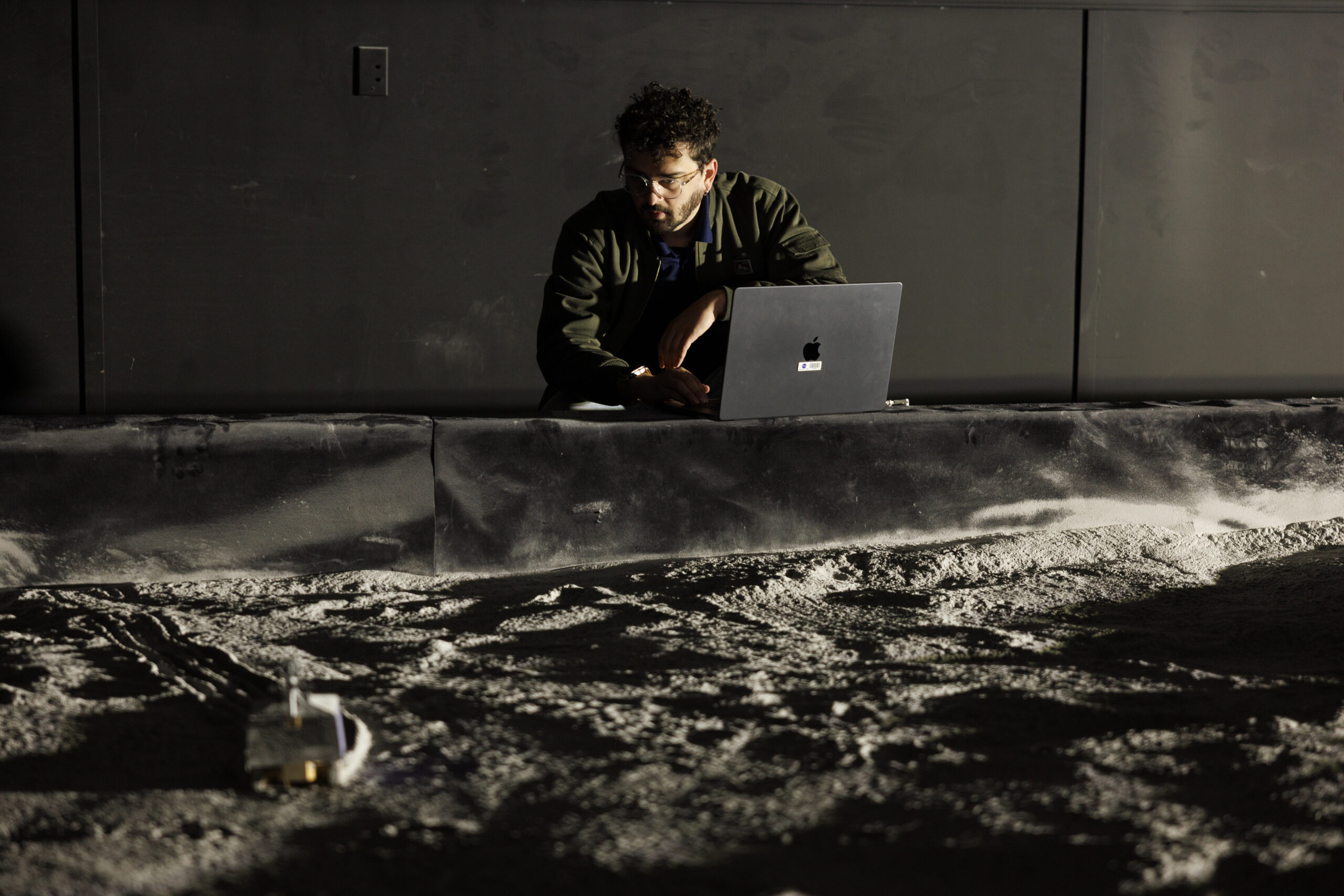 Caleb Adams kneels in a darkened room looking at a plot of sand indoors.