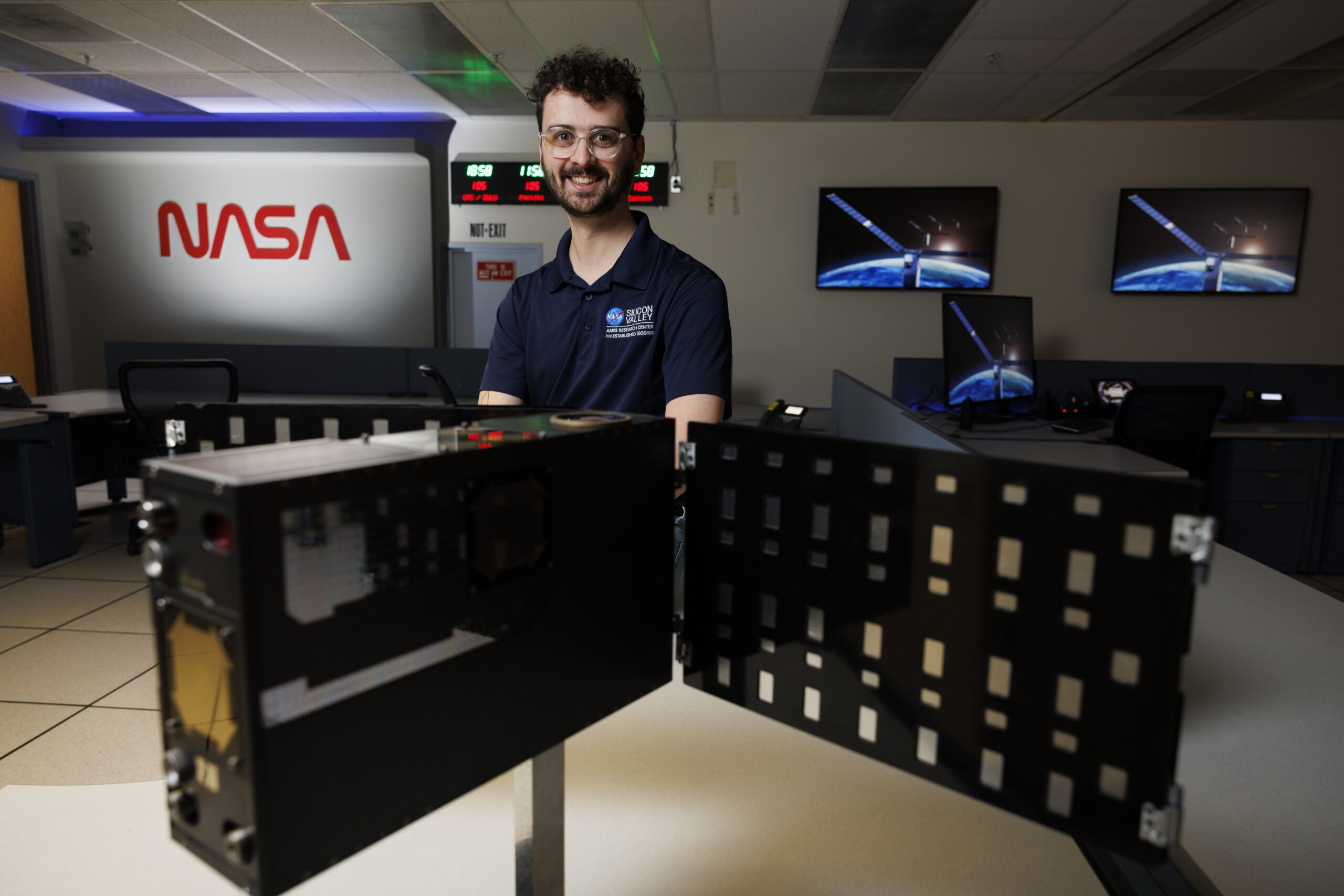 A young man in a blue polo at NASA poses behind computer equipment.
