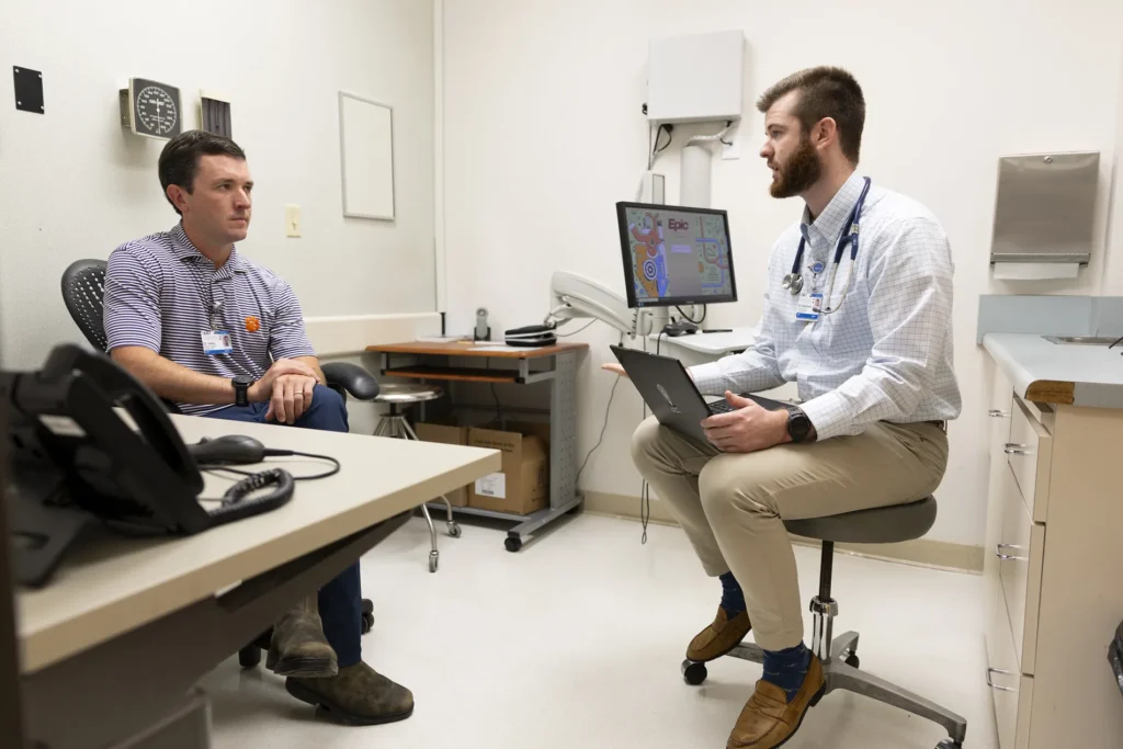 Two men sit in a doctor's office and talk.