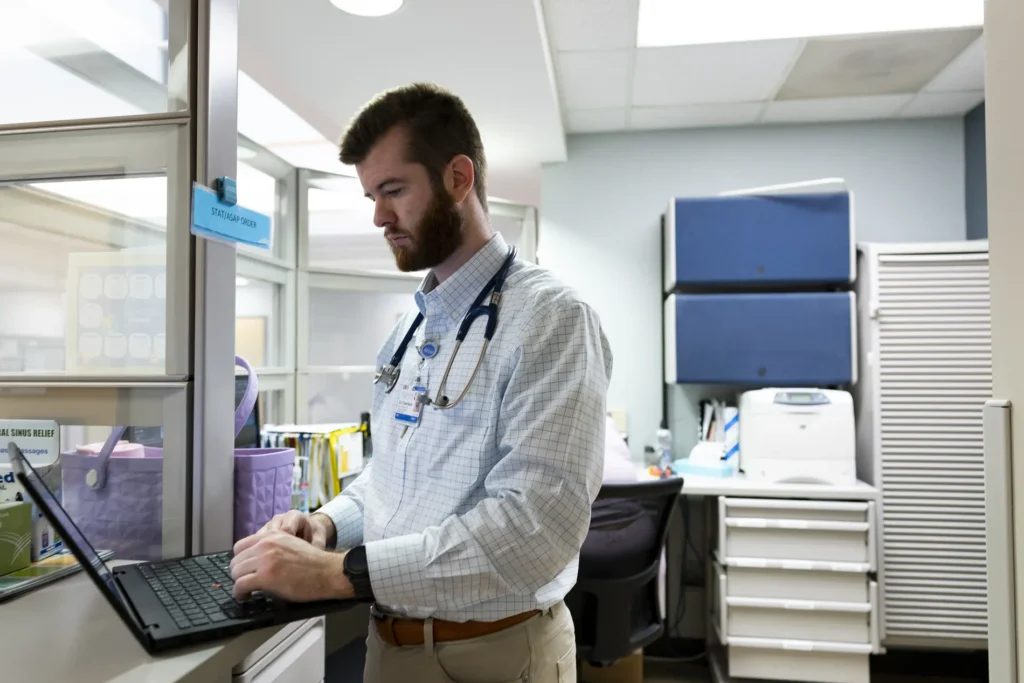 A doctor in a white checkered shirt works on a laptop.