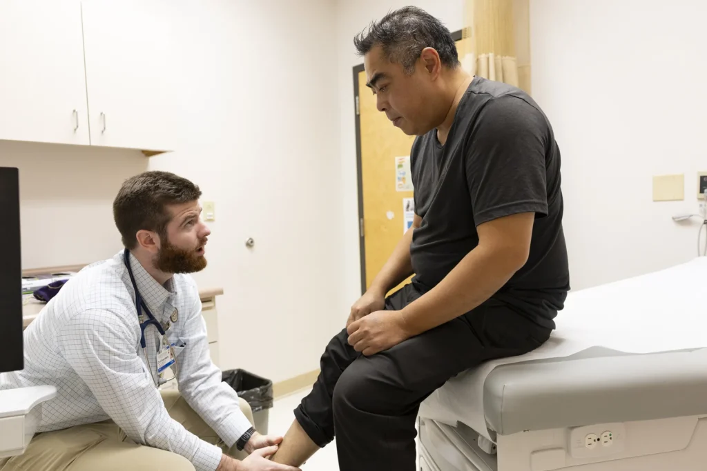 A patient dressed in black sits on an examination table while a doctor examines his leg.
