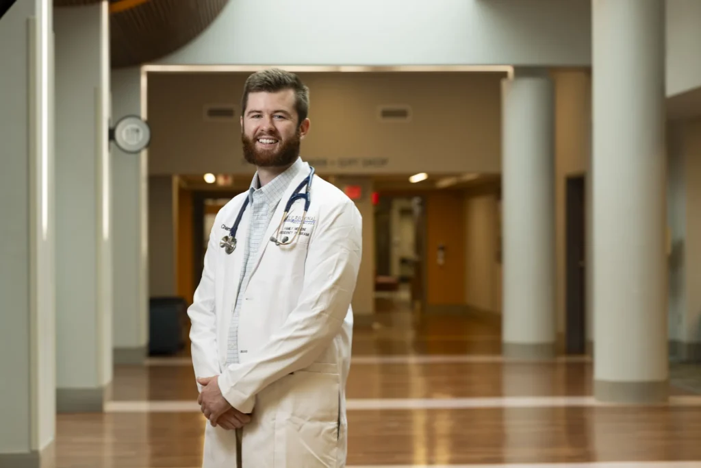 A man wearing a white medical coat poses in a brightly lit hallway.