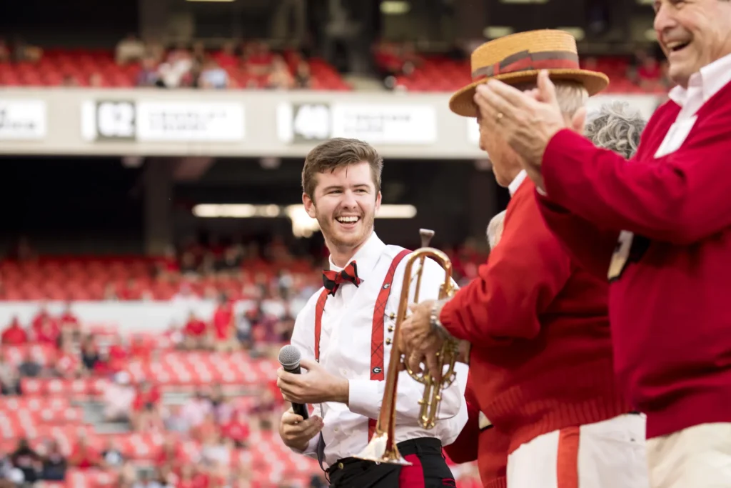 A young man in a white collared shirt and red suspenders talks with two older men in red sweaters on a football field.
