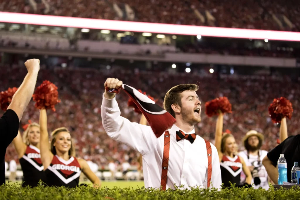 A young man in a white collared shirt and red suspenders cheers on a football field with UGA cheerleaders.