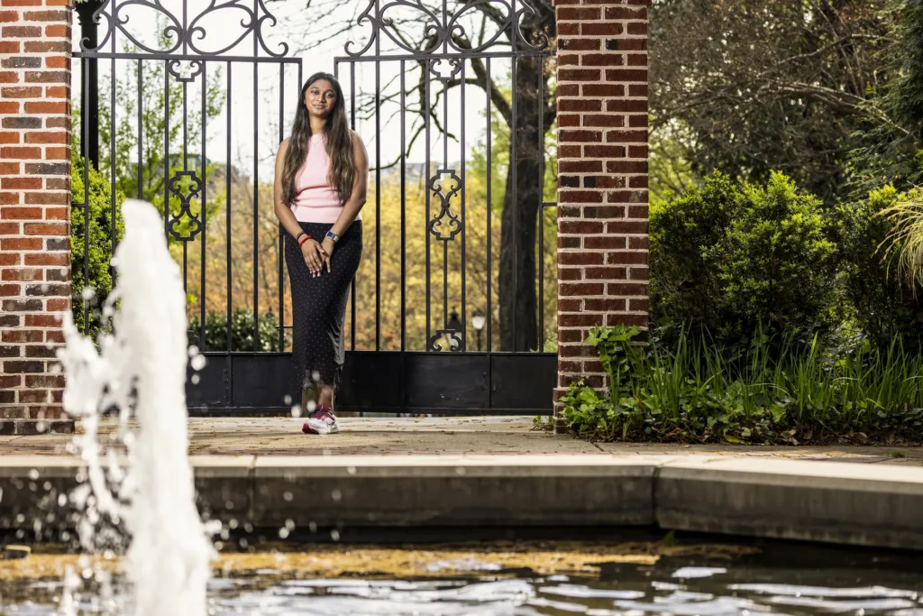 A young woman in a pink top and dark pants stands in front of an elaborate metal gate that leads to lush woodlands.