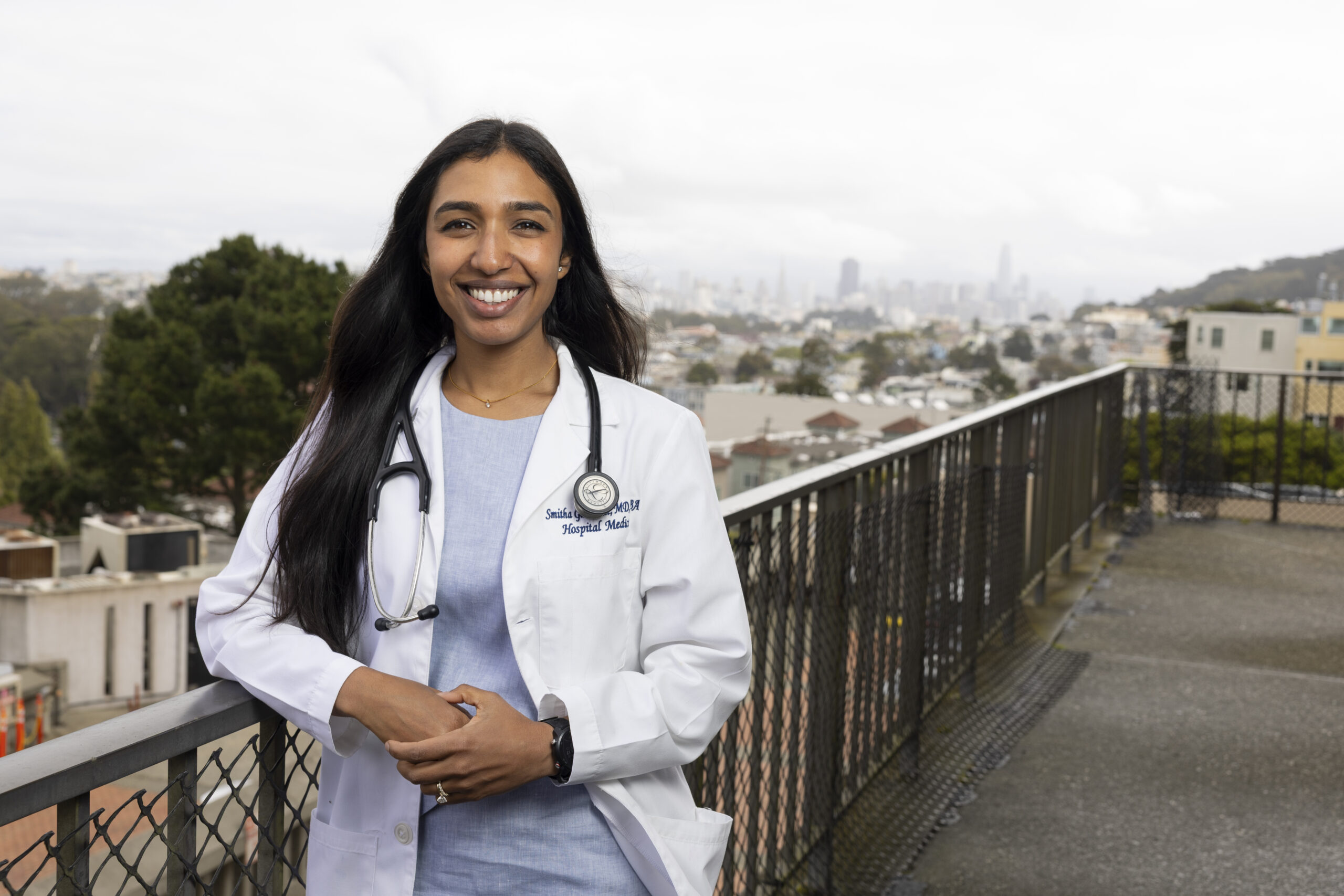 A young woman in a medical coat poses outside on a railing.