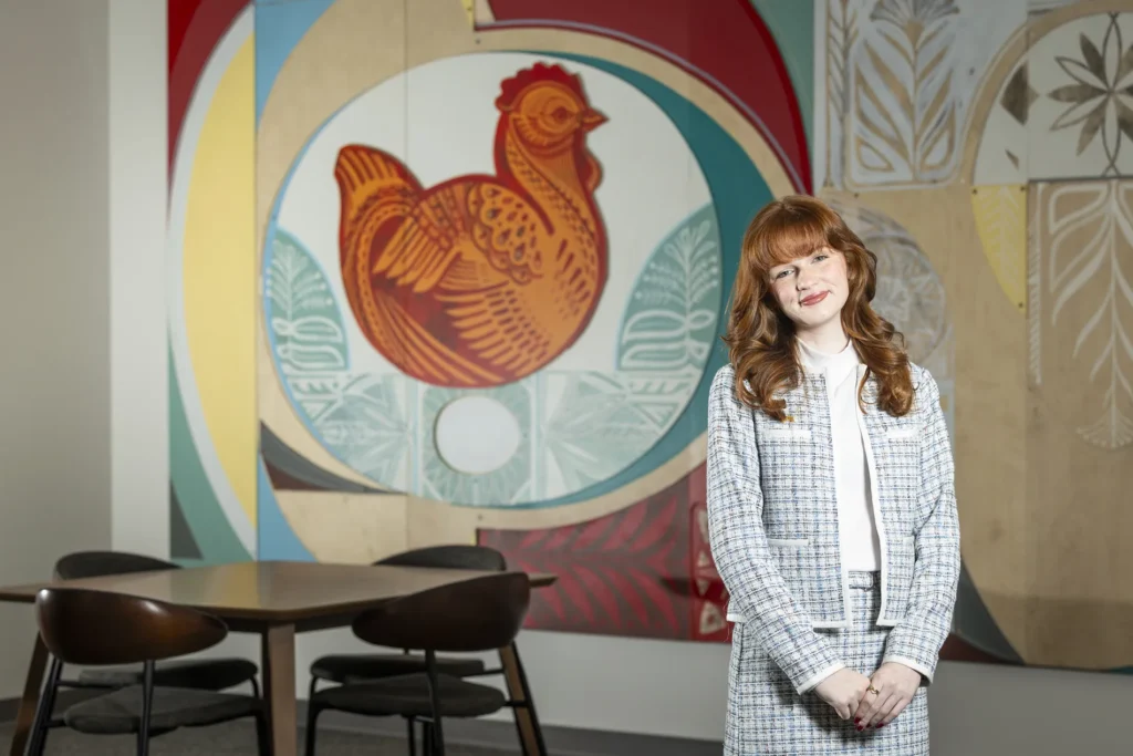 A young woman in a matching blue and white jacket and skirt stands in front of a large mural of a chicken.