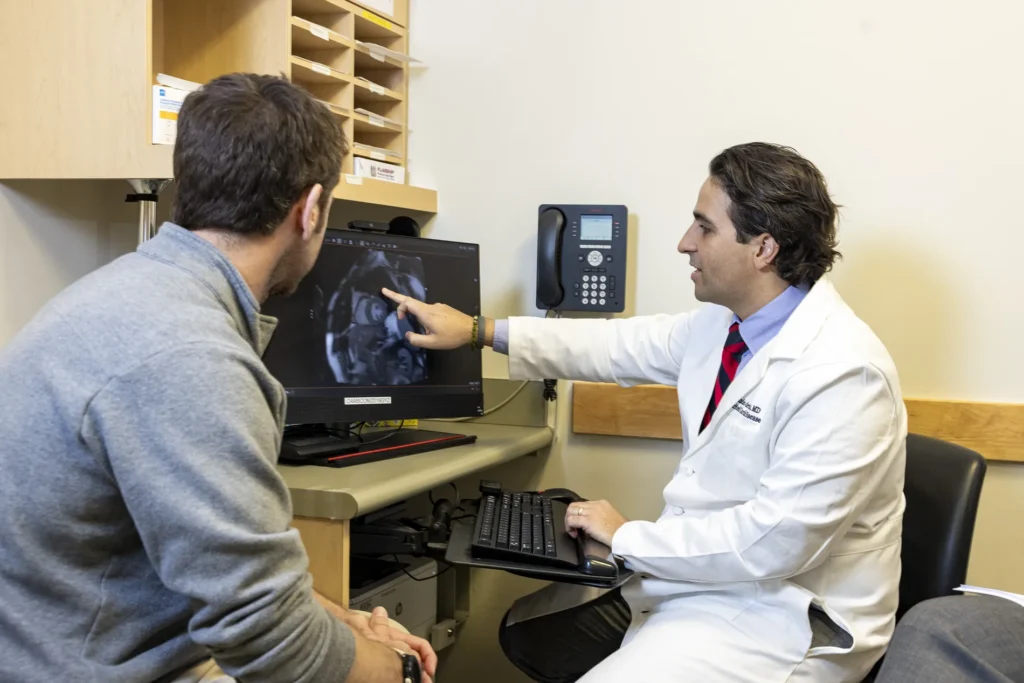 Alejandro de Feria wears a white medical coat and shows another man something on a computer.