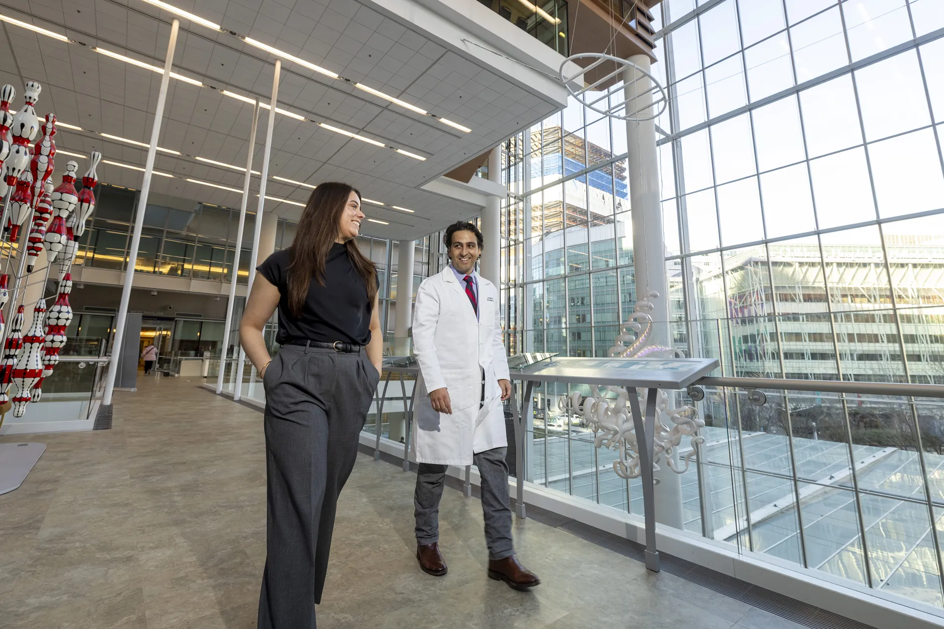 A young woman wearing dark clothing and a man in a white medical coat walk through a hallway. 
