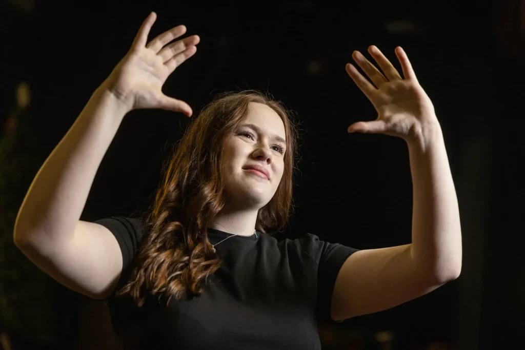 Woman on dim stage signs “imagine” in American Sign Language, hands raised near face, lit warmly against dark background.