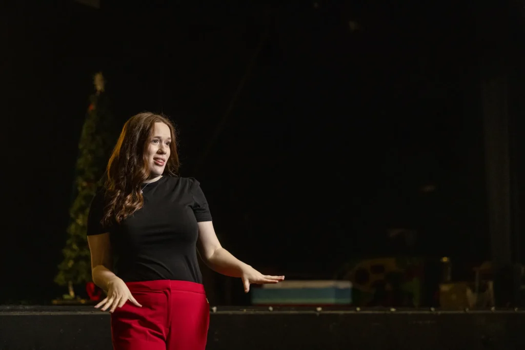 Woman in black top and red pants signs “dress” in American Sign Language on a dim stage, smiling under soft lights.