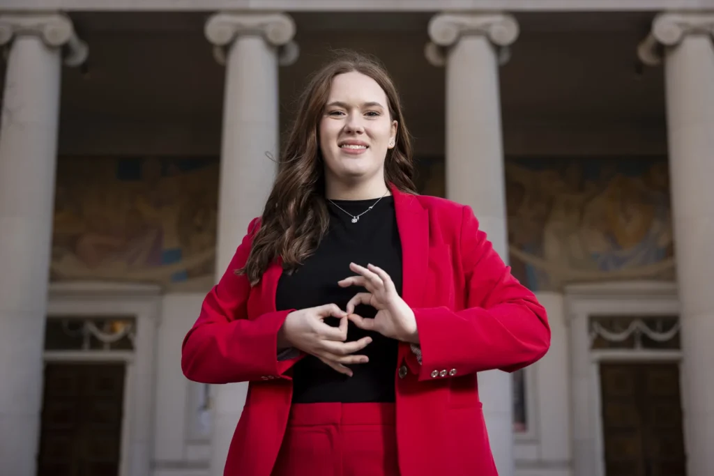 Woman in red suit standing outdoors in front of building with columns signs “interpret” in American Sign Language.