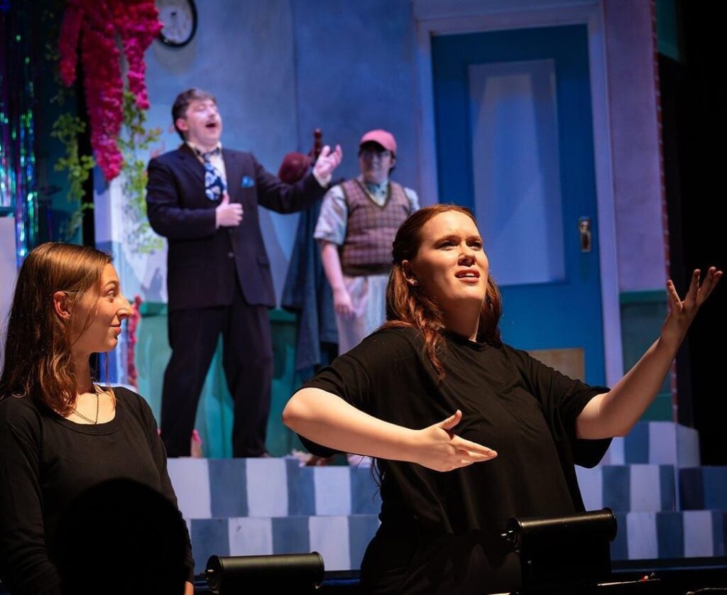 Two actors perform on stage. A sign language interpreter dressed in black clothing stands in front of the stage to translate with ASL.