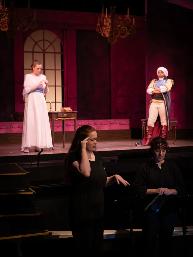 A woman in black gestures in front of actors on a stage.