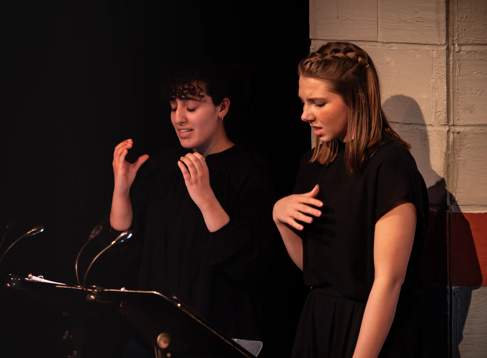 Two women dress in black clothing gesture with their hands while looking down at a script.