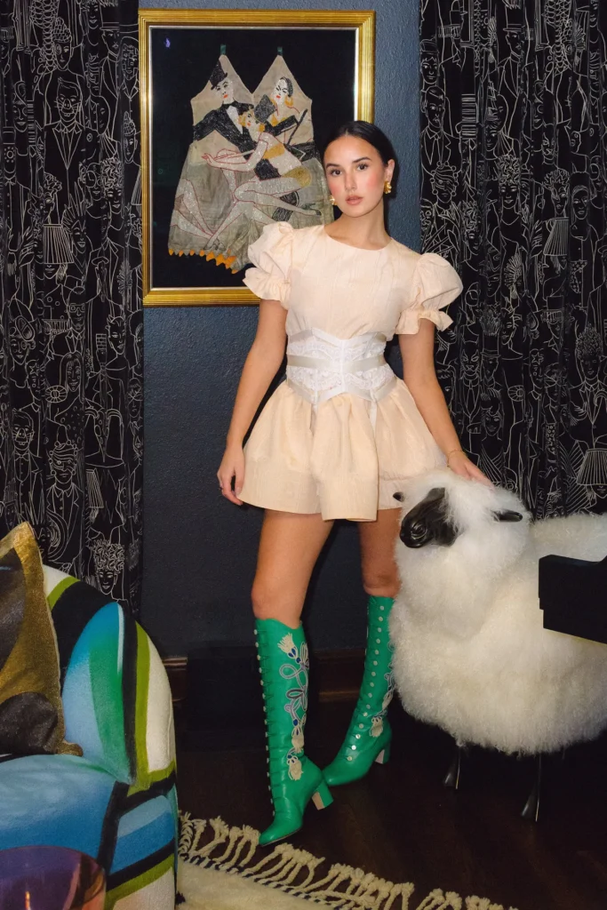 A woman in a short, pale dress and green boots poses in an eclectically decorated room.