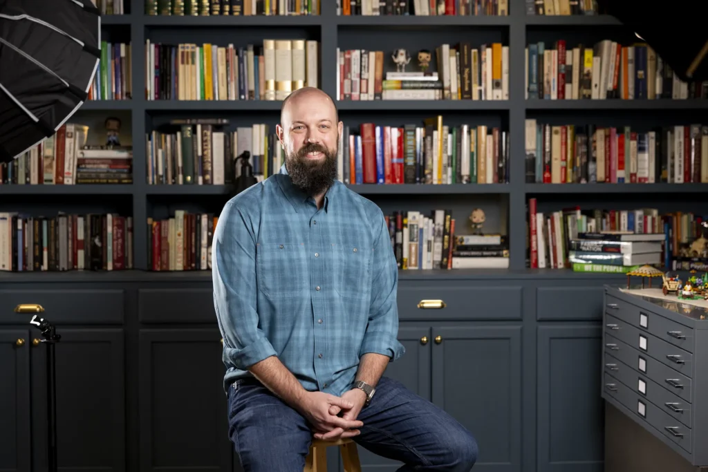 A man in a blue checkered shirt sits in front of a bookshelf.