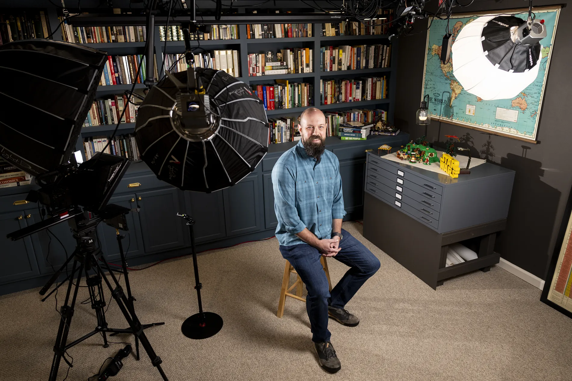 A man in a blue checkered shelf sits on a stool in front of a bookshelf and lighting equipment.