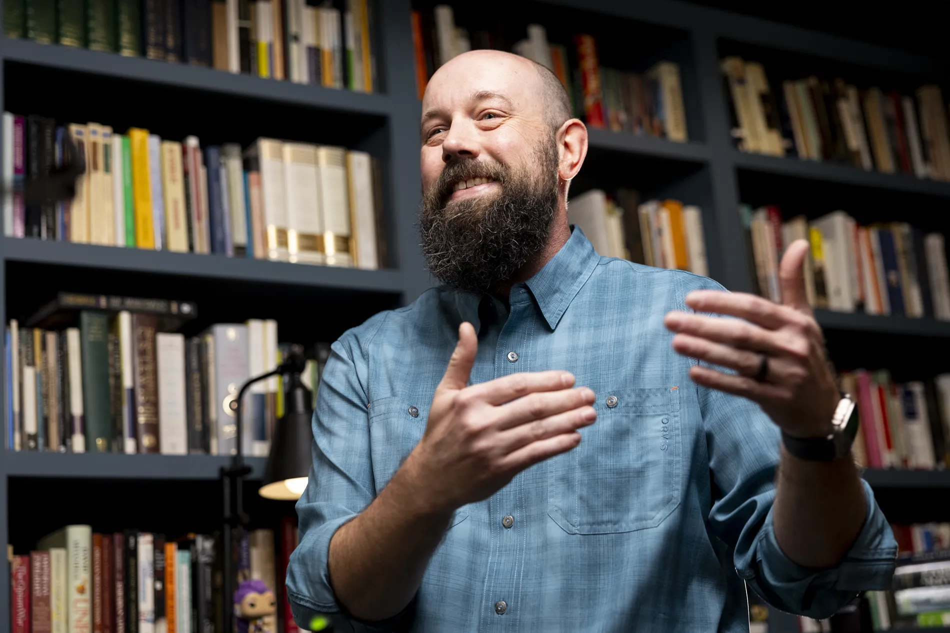 A man in a blue checkered shirt gestures as he talks in a book-filled room.
