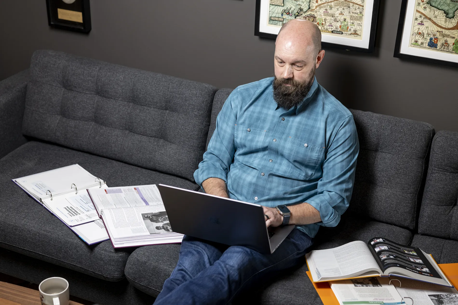A man in a blue checkered shirt sits on a couch and works on his laptop.
