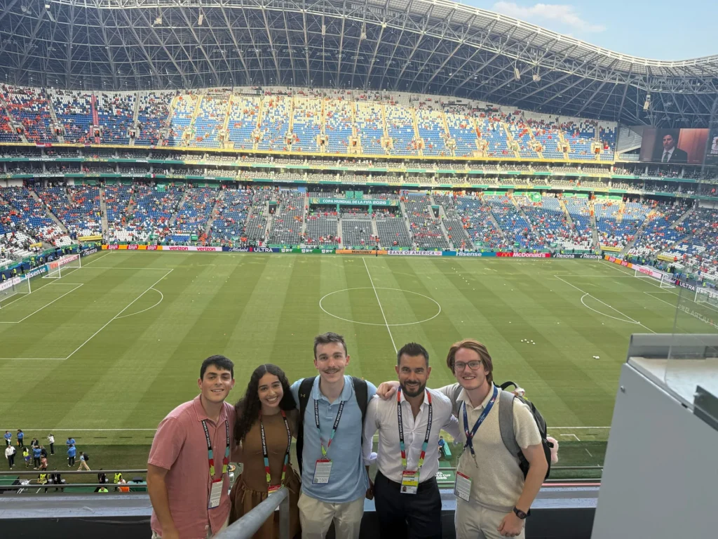 A group of five people stand in stadium bleachers with a large playing field behind them.
