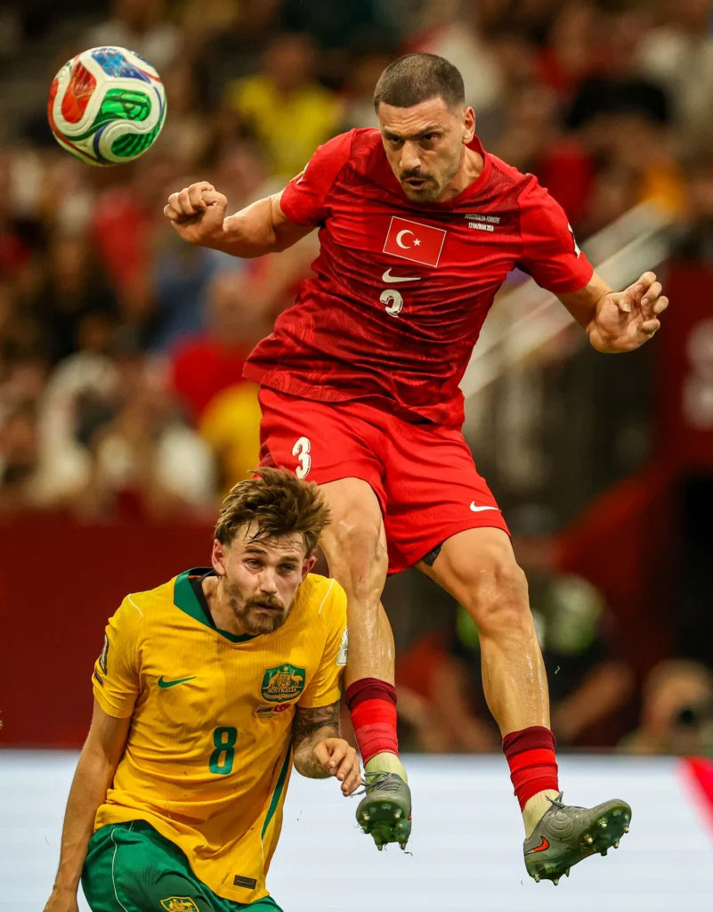 A man in a red soccer uniform jumps in the air while a man in a yellow soccer uniform stands next to his leg.