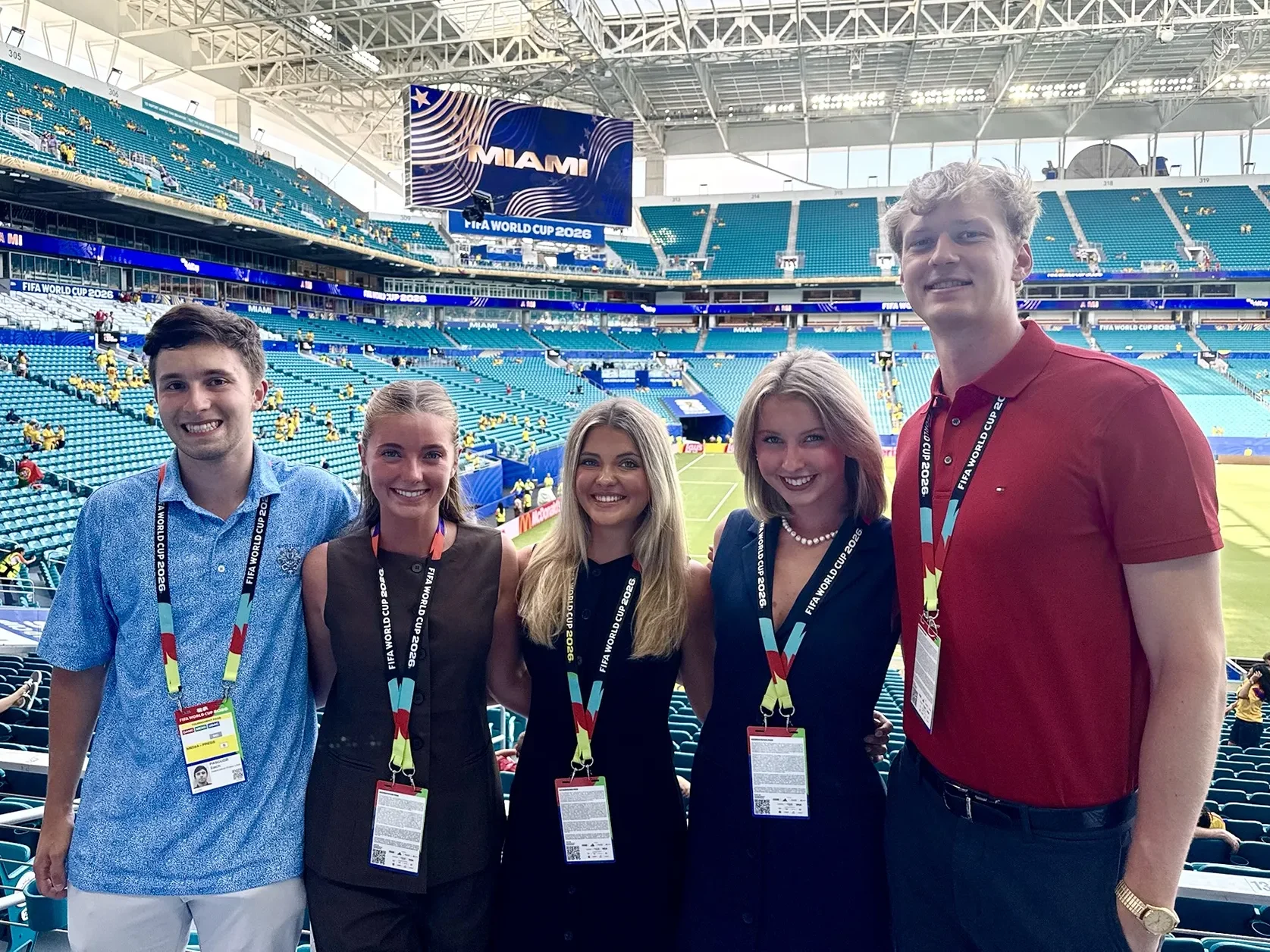 Five people wearing press badges stand in the bleachers with a large stadium behind them.