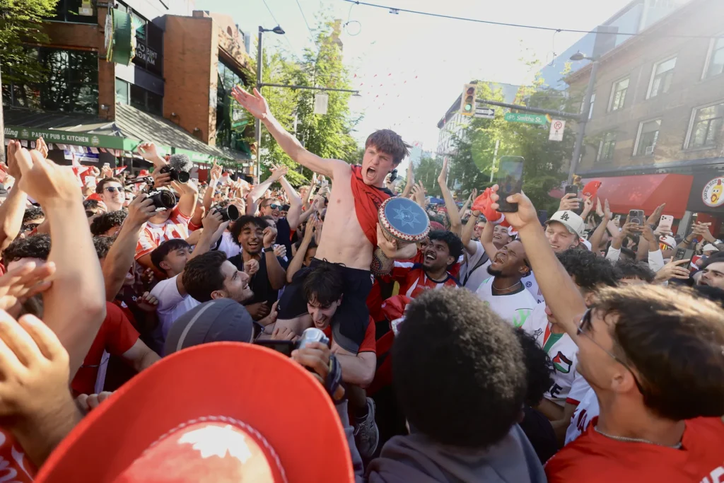 A crowd of soccer fans surround a shirtless man who is sitting on another's shoulders.