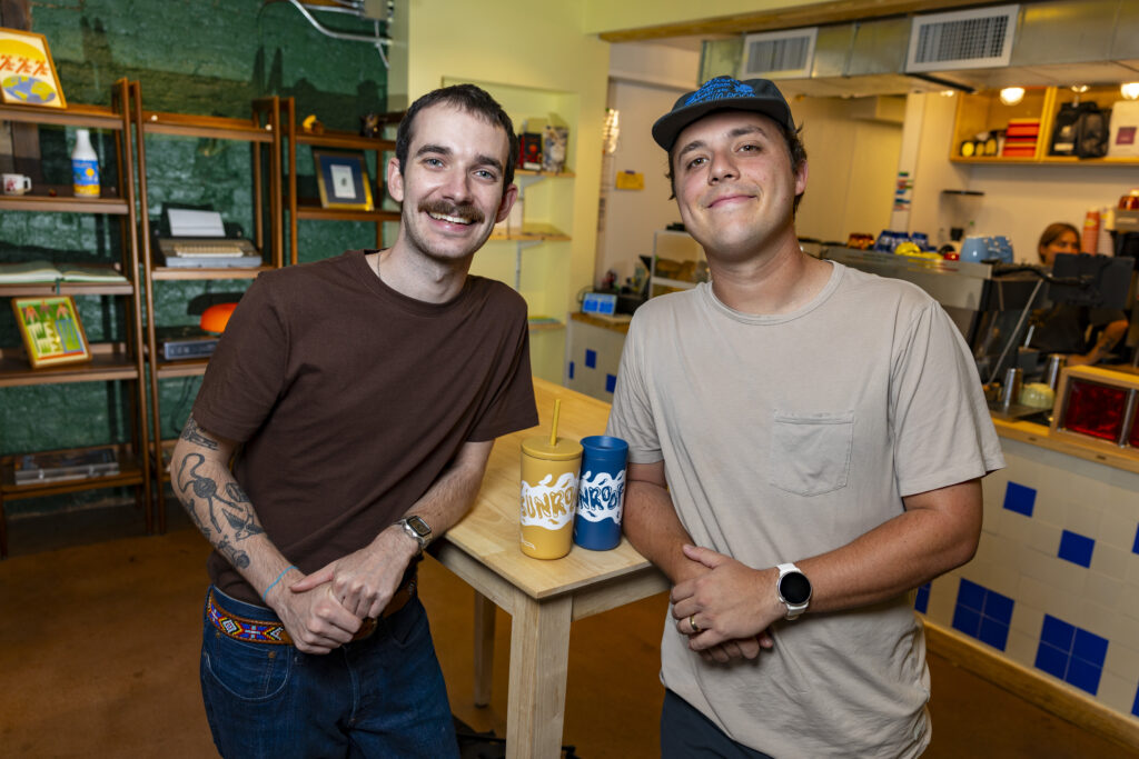 Reid Finnegan and Nick Alliston pose together in front of a tall wooden table in a cafe.