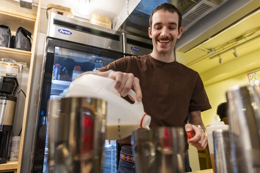A young man wearing a brown t-shirt pour milk in a cafe kitchen.