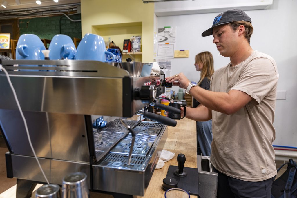 A young man wearing a dark baseball cap and a light t-shirt operates an espresso machine in a cafe.