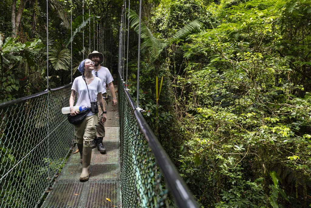 Two people wearing white t-shirts, khaki pants, and tall boots walk across a bridge surrounded by lush, green forest.