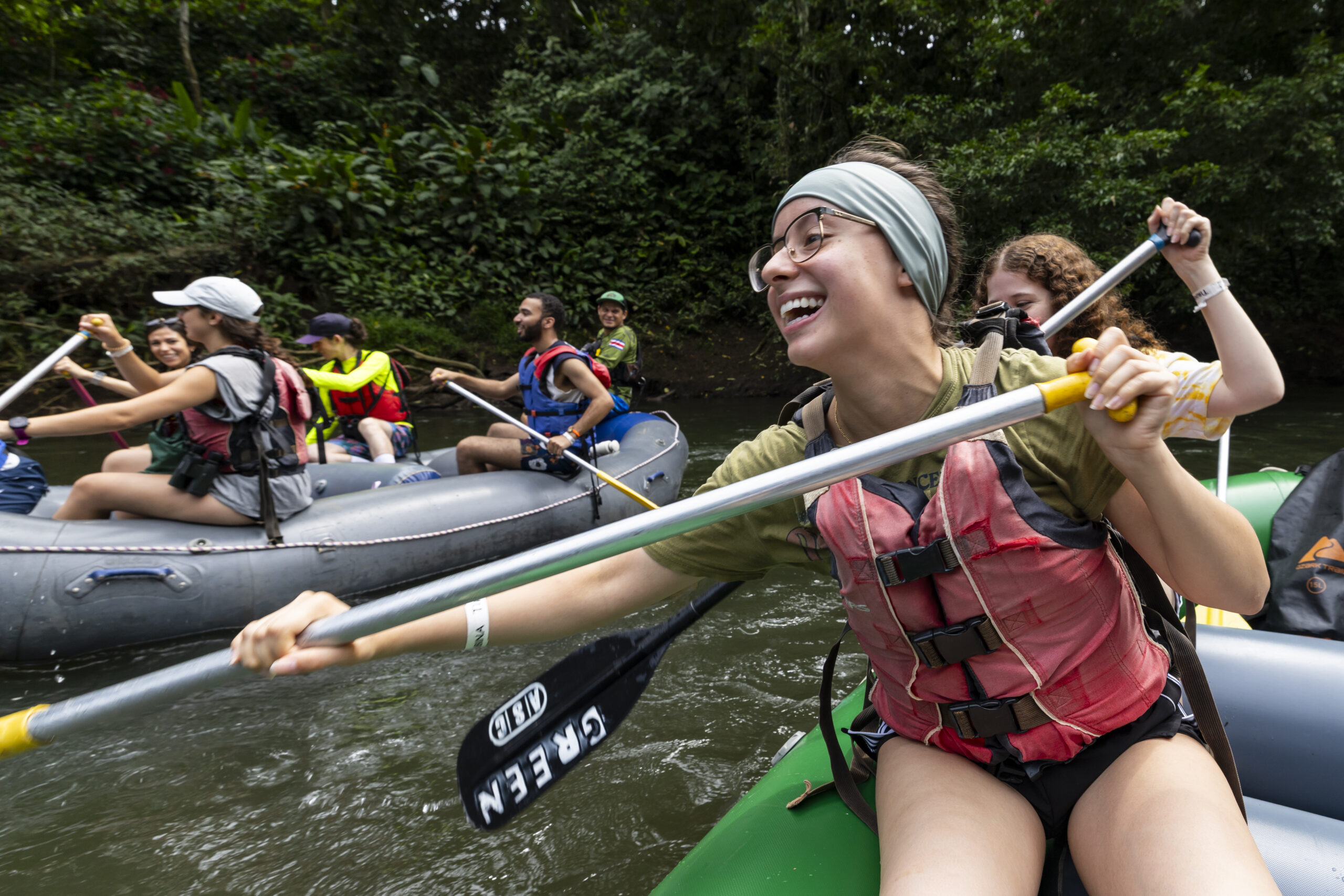 A closeup of a group of college students paddling inflatable rafts down a river.